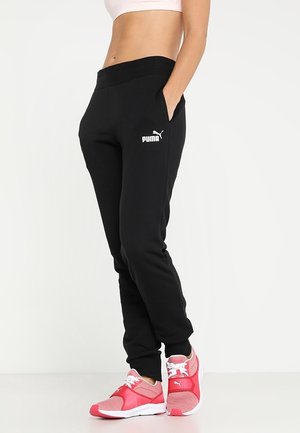 Femme portant un jogging noir Puma et des baskets roses Puma, debout les mains dans les poches devant un fond blanc uni.