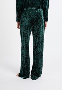 Zelené sametové kalhoty širokého střihu s jemnou texturou a lesklým povrchem. Mají elastický pas a jemné vzory po celém povrchu.