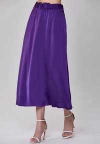 Jupe midi en satin violet avec une taille ceinturée, texture lisse, design fluide et ourlet qui atteint le mollet. Associée à des sandales blanches.