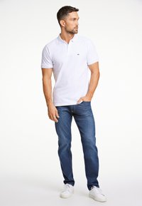 Valkoinen polo-paita lyhyillä hihoilla, musta logo, yhdistetty tummansinsisiin farkkuihin ja valkoisiin lenkkareihin. Yksinkertainen tausta, rento tyyli.