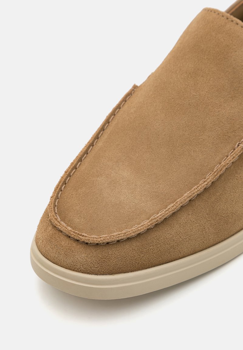 Zapato sin cordones de ante marrón, con puntera redondeada, costura decorativa a lo largo del borde y una suela de goma de color claro.