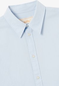 Hellblaues Button-up-Hemd aus Baumwolle, klassischer Kragen, sieben Knöpfe, feine Textur, kleines Markenlabel im Kragenbereich.