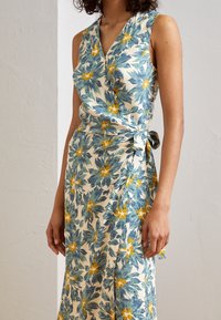 Kukkakirjailtu muhkean kankaan mekko, jossa on vihreitä lehtiä ja keltaisia kukkia vaalean kermanvärisessä taustassa, ja vyötäröllä solmittava yksityiskohta.