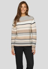 Frau in einem gestreiften Langarmpullover in Beige, Weiß, Grau und Schwarz sowie einer schwarzen Hose, die vor einem schlichten grauen Hintergrund steht.