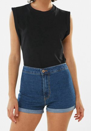 Persoon draagt een mouwloze zwarte top, gestopt in een high-waisted blauwe spijkershort met opgerolde zoom en een knoopsluiting aan de voorkant.