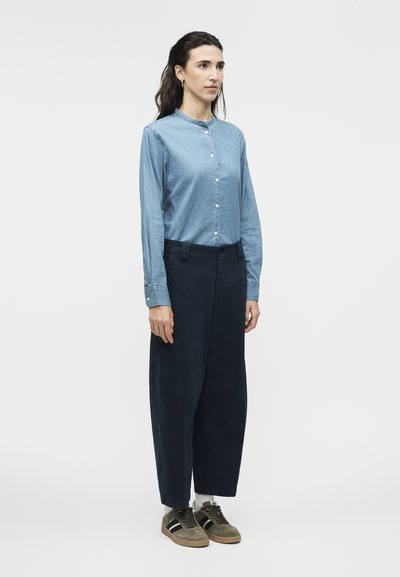 Γυναίκα που στέκεται φορώντας ανοιχτό μπλε πουκάμισο με κουμπιά, σκούρα παντελόνια με φαρδιά μπατζάκια, λευκές κάλτσες και πράσινα αθλητικά παπούτσια με μαύρες και λευκές ρίγες.