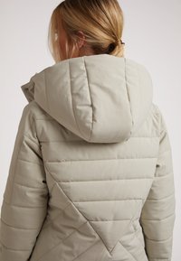 Lys beige dunjakke med hætte. Har diagonalt quiltet mønster på ryggen, blød tekstur og en struktureret hætdesign.