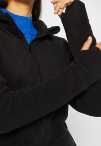 Mustat fleece-takit, joissa on vetoketju edessä ja korkea kaulus; pitkähihaiset hihat, joissa on peukalonreiät, ja teksturoitu sininen sisäkerros.