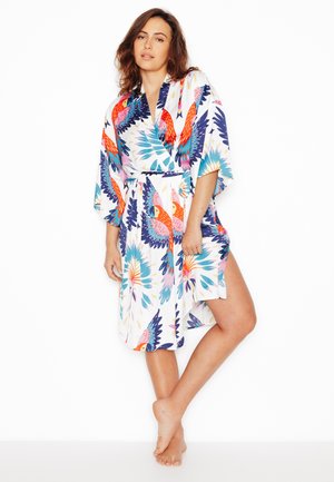 Femme debout pieds nus portant une robe portefeuille blanche avec des motifs vibrants de perroquets colorés et de plumes.