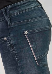 Jeans en denim bleu foncé avec une coupe ajustée, présentant des poches visibles, des détails en métal argenté et des surpiqûres contrastées le long des coutures.