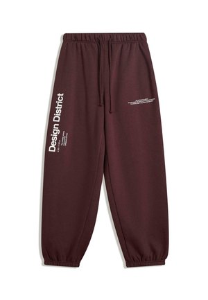 Pantaloni di felpa marrone scuro con vita ed elastici sulle caviglie, che presentano il testo bianco "Design District" sulla gamba sinistra e un testo più piccolo sulla coscia destra.