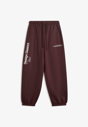 Pantaloni di felpa marrone scuro con vita ed elastici sulle caviglie, che presentano il testo bianco "Design District" sulla gamba sinistra e un testo più piccolo sulla coscia destra.