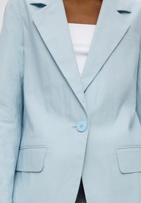 Blazer azzurro chiaro realizzato in un tessuto texturizzato, con chiusura a bottone singolo, rever affilati e due tasche frontali.