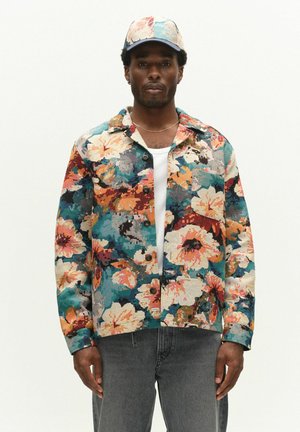 Homme portant une veste boutonnée à motifs floraux multicolores et une casquette assortie sur une chemise blanche et un jean gris, debout devant un fond uni.