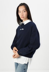 Marineblauwe sweatshirt met geborduurd logo, over een wit overhemd met kraag. Kenmerkt zich door een losse pasvorm, lange mouwen en geribde manchetten. Lichtblauwe jeans.