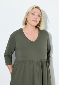 Oliwkowa sukienka z rękawami 3/4, wycięciem w serek i marszczoną talią. Wykonana z miękkiego, lekkiego materiału o luźnym kroju. Bez wzorów ani akcentów.