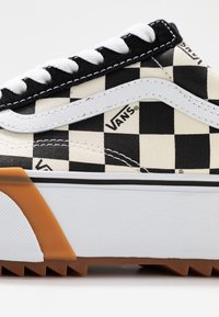 Gros plan sur une basket Vans avec un motif à damier noir et blanc, une bande en cuir blanc, des lacets blancs et une semelle en caoutchouc beige.