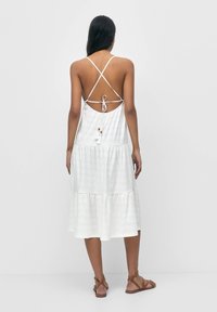 Bílé, bezramínkové šaty s vrstveným designem. Mají křížové zadní popruhy, pásek v pase a pro textura očkovaný vzor. Nosené s hnědými sandály.