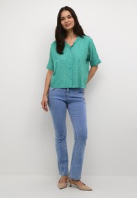 Blusa corta teal con maniche corte e pois bianchi, con bottoni davanti; abbinata a jeans azzurri chiari e scarpe col tacco nude.