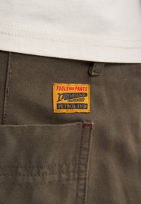 Primo piano di una tasca dei pantaloni marroni con una toppa gialla etichettata "Strumenti e Parti Campionato Lumberjack Petrol Ind" sotto l'orlo di una camicia bianca.