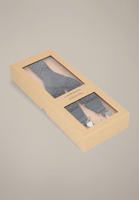 Czarne szelki w białe kropki, wyposażone w srebrne metalowe klipsy, zapakowane w przezroczyste prostokątne pudełko z brązowym zewnętrzem z papieru kraft.