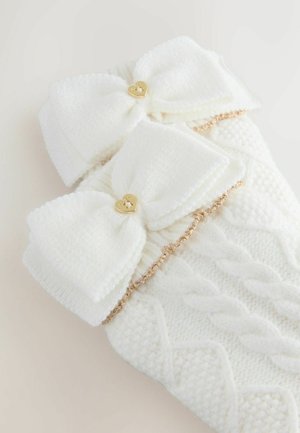 Valkoiset neulotut säärystimet, joissa on kaksi suurta nauhaa kullanvärisillä sydänkoristeilla ja kultaketjuyksityiskohdalla, esittelevät teksturoitua kaapelikuviota.