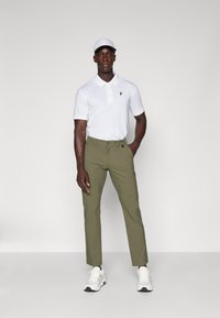 Hvid polo shirt med et sort logo, olivengrønne bukser og hvide sneakers med sorte detaljer. Modellen står foran en ensfarvet baggrund.