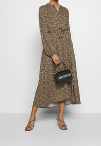 Abito lungo a maniche lunghe di colore marrone con motivo floreale, con vita legata, che arriva a metà polpaccio. La modella tiene in mano una piccola borsa nera e indossa sandali con strappi.