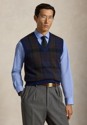 Homme portant des lunettes rondes, une chemise bleue, une cravate sombre, un gilet en tricot à carreaux et un pantalon gris plissé, les mains dans les poches, sur un fond uni.