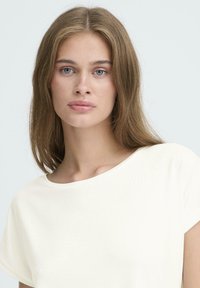 Ung kvinde med langt lysbrunt hår og blå øjne iført en helt hvid t-shirt med rund hals mod en neutral baggrund.