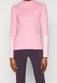 Różowy top sportowy z wysokim kołnierzem i zamkiem na połowie długości. Posiada białe trzy paski na rękawach i małe logo na piersi.