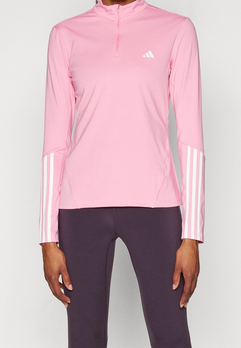 Różowy top sportowy z wysokim kołnierzem i zamkiem na połowie długości. Posiada białe trzy paski na rękawach i małe logo na piersi.
