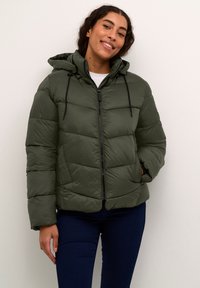 Doudoune en vert foncé avec fermeture éclair, design matelassé, capuche ajustable et poignets élastiques. Portée avec un jean noir.