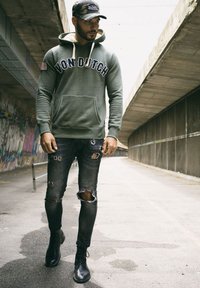 Housse verte avec le logo "Von Dutch", poche avant et capuche avec cordon de serrage ; associée à un jean noir déchiré et des bottes noires à lacets.