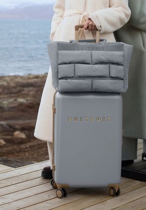 Person in light coat holding beige suitcase handle with gray puffy bag on top, standing on wooden deck near rocky shore and water.