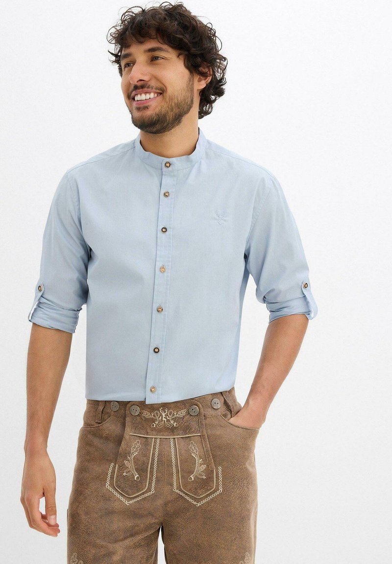 Camicia azzurro chiaro con colletto alla mandarino, dotata di bottoni marroni e maniche arrotolate. Indossata con lederhosen marroni ricamati.