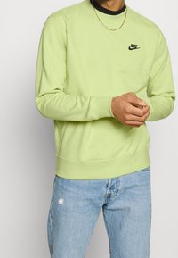 Sweatshirt vert clair avec des poignets et un ourlet côtelés, arborant un petit logo Nike noir sur la poitrine, porté avec un jean bleu déchiré.