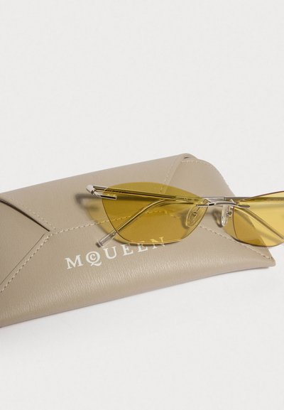 Keltaiset aurinkolasit langarunkoilla lepävät beigeissä nahkakuorissa, joissa on pistetetäytteinen kuvio ja valkoisilla kirjaimilla kirjoitettu sana "McQueen".