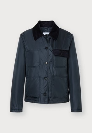 Jeune femme portant une veste boutonnée bleu foncé avec des poches, un pantalon noir et des bottes noires, debout face à un fond uni.