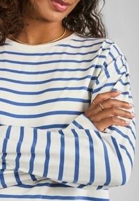 Camicia a maniche lunghe a righe bianche e blu realizzata in cotone morbido, con scollatura a girocollo e vestibilità rilassata. La mano del modello mostra anelli delicati.