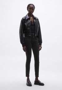 Chaqueta bomber de cuero negro con puños acanalados, combinada con vaqueros ajustados negros y mocasines. El modelo está de pie frente a un fondo claro y liso.