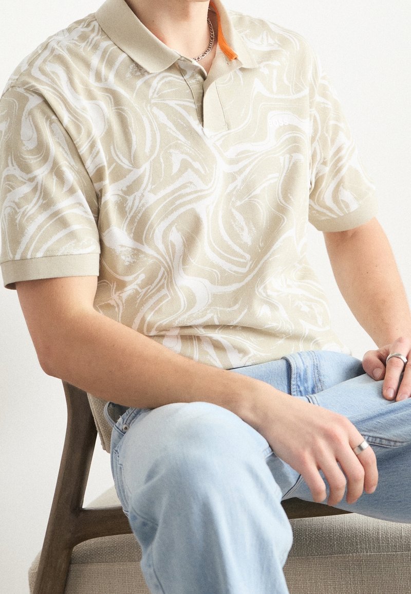 Homme assis sur une chaise en bois porte un polo beige et blanc tourbillonnant et un jean bleu clair, avec des bagues en argent aux doigts.