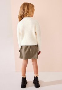 Weißer, flauschiger Pullover, olivgrüner Rock, schwarze Stiefeletten mit Reißverschlüssen und hellfarbige Socken. Weicher Stoff und lockere Passform.