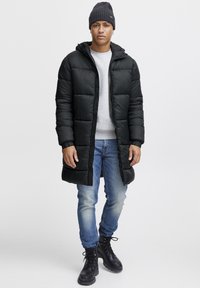 Abrigo largo acolchado negro con capucha sobre un sudadera gris, combinado con jeans slim azules y botas negras con cordones. Textura suave en todo.