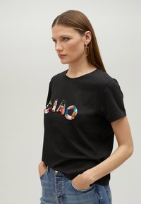 Svart t-shirt med kort ärm som har ett flerfärgat paljetttryck med texten "CIAO". Tillverkad av mjuk bomull med normal passform och rund halsringning.