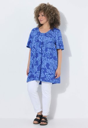 Frau mit lockigem Haar, die eine blaue gemusterte Tunika, weiße Hose und schwarze Sandalen trägt und vor einem weißen Hintergrund steht.