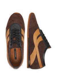 Brūni zamšādas sporta apavi ar gaiši brūniem akcentiem, melniem dekoratīviem elementiem un strukturētām šņorēm. Iekļauj gludu gumijas zoli un logo etiķeti iekšpusē.