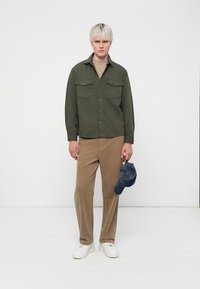 Green button-up shirt with two chest pockets, tan corduroy pants, and a blue cap featuring a textured pattern. White sneakers complete the look.