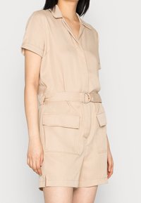 Tutina corta beige realizzata in tessuto leggero, con collo a camicia, maniche corte, vita con cintura e ampi tasconi frontali.