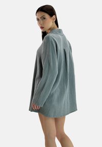 Oversized, gestructureerde overhemdjurk in gedempte teal, met lange mouwen, een kraag en een subtiel geplooid detail aan de achterkant.